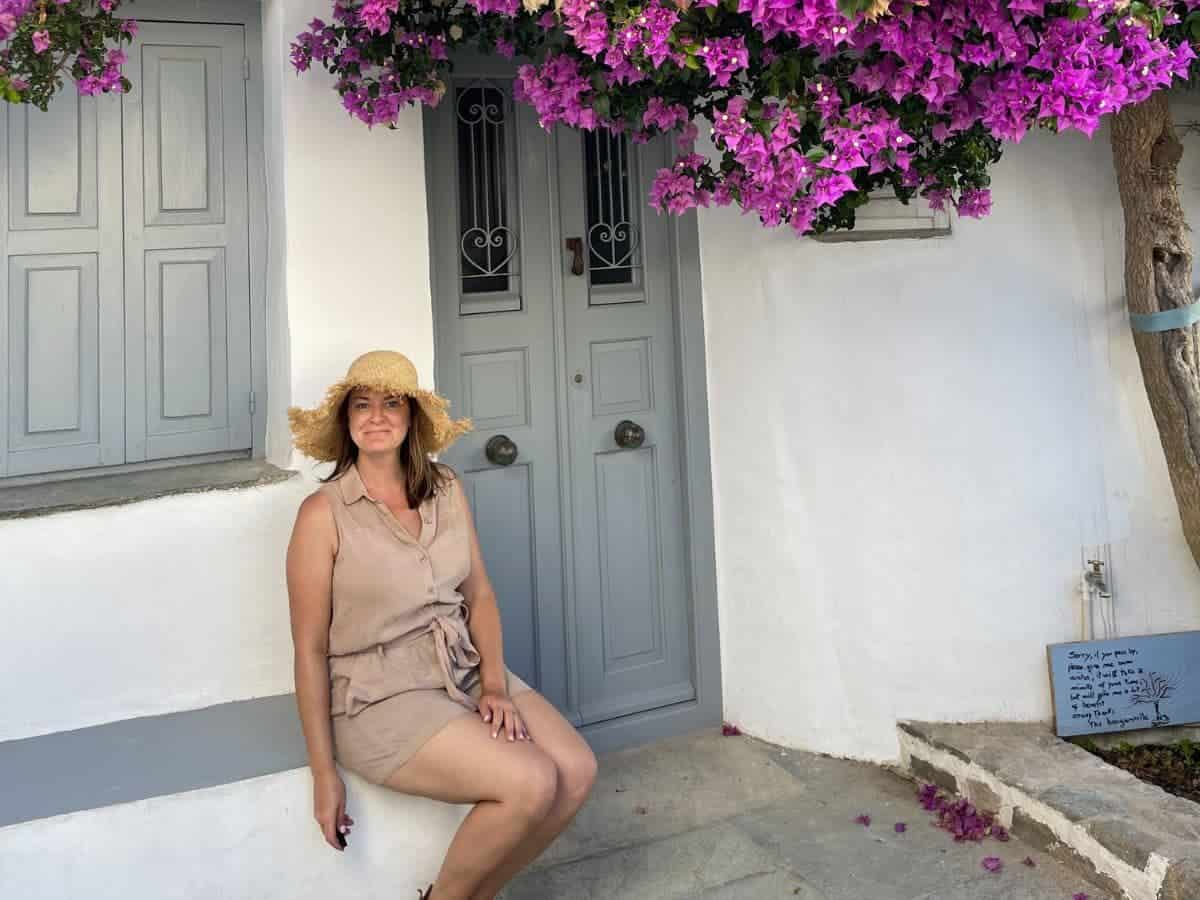 Smiling woman enjoying solo travel in Greece, sitting casually on steps by a traditional Greek house with a gray door, framed by vibrant bougainvillea
