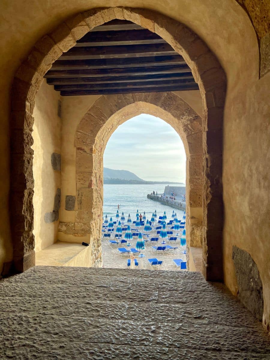 A scenic view of Cefalù Beach in Sicily, framed by an ancient stone archway. The weathered stone passageway opens up to a beach filled with neatly arranged blue umbrellas and sun loungers, with the calm sea and a distant pier in the background. The image captures a blend of historic architecture and coastal charm.