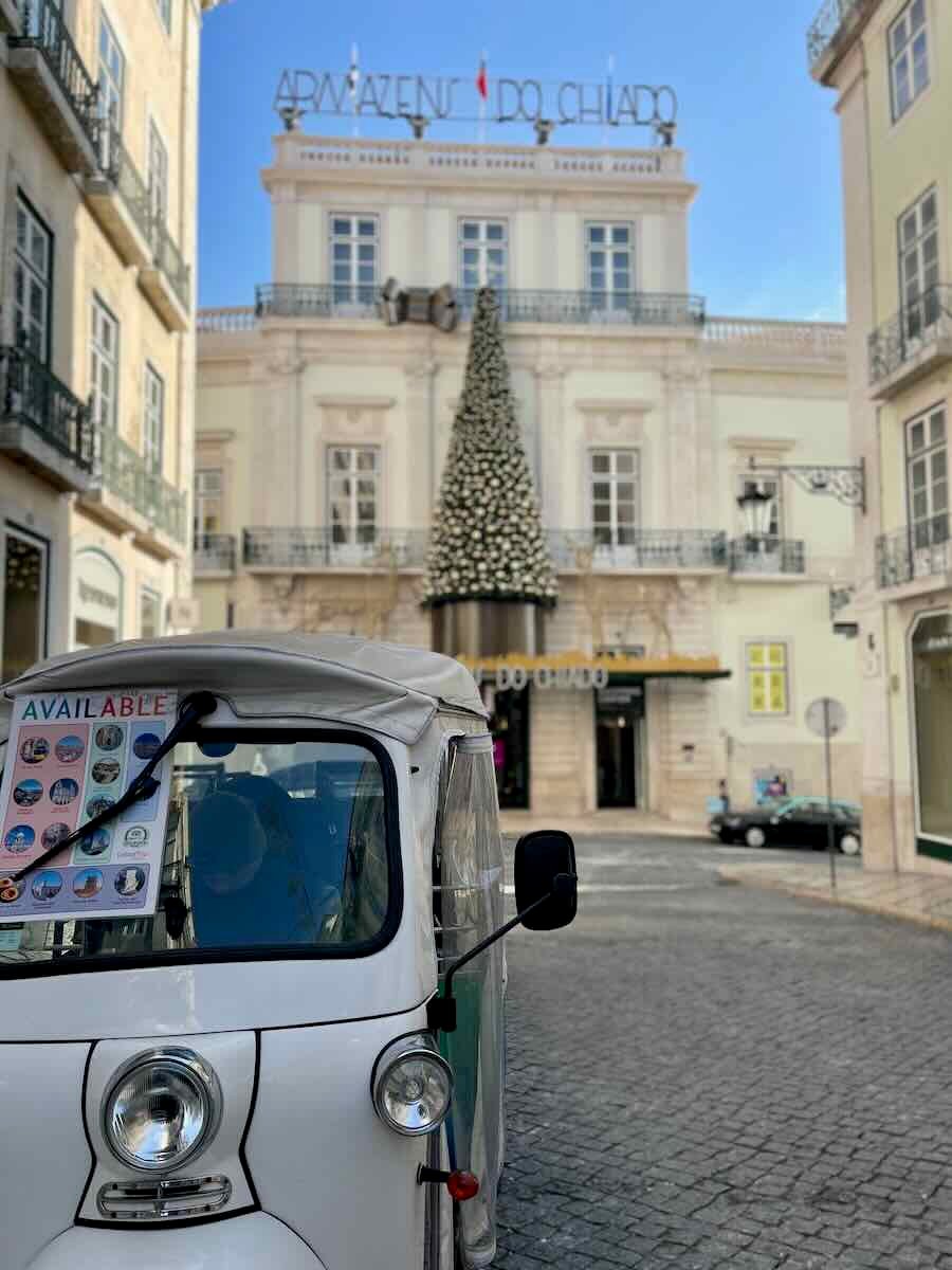 A white tuk-tuk vehicle parked in front of the Armazéns do Chiado shopping center in Lisbon, Portugal, which is adorned with a large, decorated Christmas tree in front of the building. The scene is set on a clear day, with the historic architecture of the city in view.