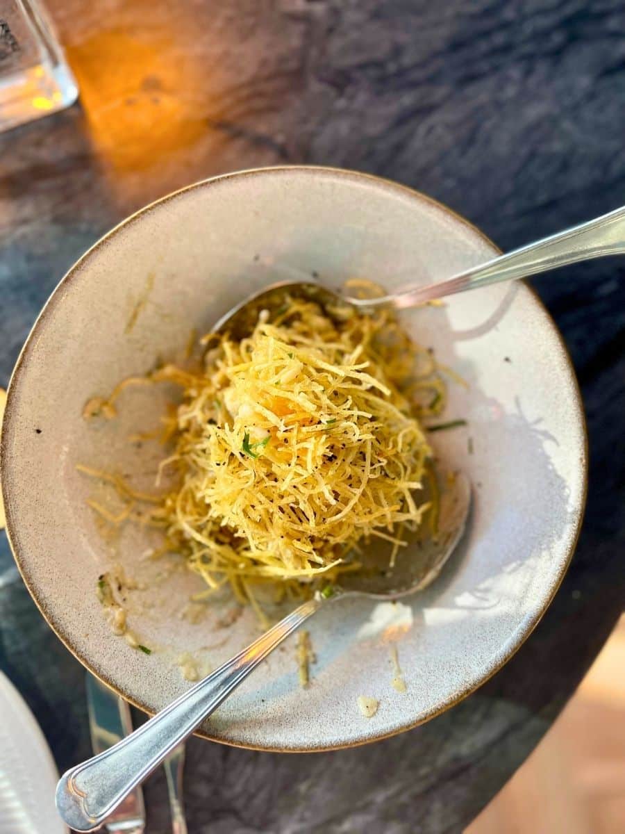 A beautifully plated dish featuring crispy, golden potato strands delicately piled atop a savory mixture, served in a rustic ceramic bowl. Two silver spoons rest on the dish, suggesting it is meant for sharing. The background features a dark, textured tabletop with warm ambient lighting casting a soft glow, enhancing the dish's appeal. The crispy texture and vibrant golden hues of the potatoes contrast with the smooth, neutral tones of the bowl, making for an inviting and visually appealing presentation.