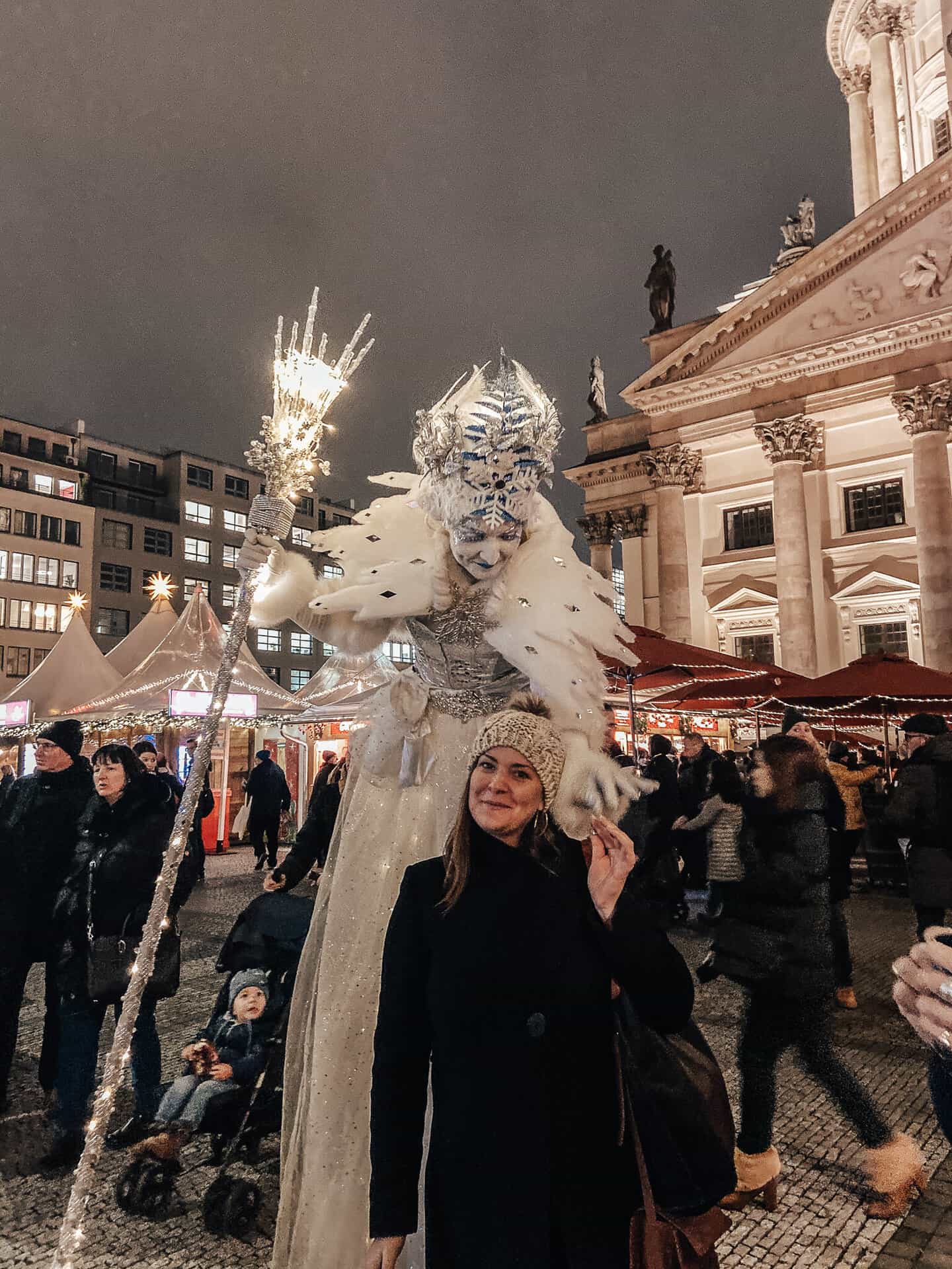 A woman poses with a performer dressed as an elaborate winter queen, complete with a sparkling crown and staff, at a festive outdoor market. The scene is set in front of a grand, illuminated building, with market stalls and a crowd of people in the background, creating a lively and magical holiday atmosphere.