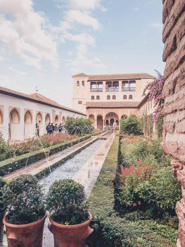 Inside Alhambra. Elegant courtyard garden with a central reflective pool flanked by lush greenery and trimmed hedges. The pool is lined with small water fountains, creating a serene atmosphere. In the background, an arched building with a terracotta roof and a series of windows overlooks the garden. 